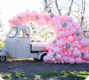 a pink and silver balloon arch incorporating a vintage three wheeled truck in a sunny outdoor setting