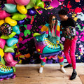 Dos personas vestidas con ropa de colores sonríen y posan con un globo de 27" SuperShape 90s Party Inline Roller Skate Foil Balloon de Anagram/MD y globos SO FLY delante de un fondo de pintura de neón, de pie sobre un suelo de madera.