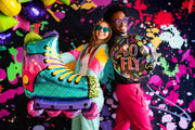 Dos personas con coloridos trajes retro y gafas de sol posan delante de un fondo de pintura salpicada. Una de ellas sostiene un globo Anagram/MD 27" SuperShape 90s Party Inline Roller Skate Foil Balloon; la otra sostiene un globo redondo "SO FLY".
