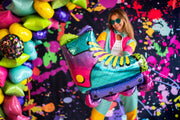 Una mujer con un colorido atuendo y gafas de sol sostiene el Globo de Foil Anagram/MD 27" SuperShape 90s Party Inline Roller Skate delante de un fondo de pintura salpicada con brillantes globos en forma de estrella, creando una escena lúdica y festiva.