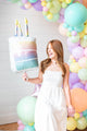 A woman in a white dress smiles while holding the 28" Holographic SuperShape Pearlized Birthday Foil Balloon by Anagram/MD in front of a colorful pastel balloon arch decorated for a birthday celebration.
