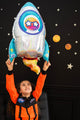 Un enfant vêtu d'une combinaison d'astronaute orange sourit en tenant un ballon en feuille Anagram/MD 29" Holographic SuperShape Outer Space. Derrière eux, un mur noir décoré d'étoiles et de planètes crée une toile de fond amusante sur le thème de l'espace.