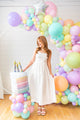 A woman in a white dress smiles beside an Anagram/MD 28" Holographic SuperShape Pearlized Birthday Foil Balloon, with pastel party decor and a faux rainbow cake, in a bright room with wood floors and white walls.
