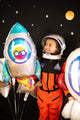 Un jeune enfant rayonne en tenant le ballon en aluminium SuperShape Outer Space de 29 pouces d'Anagram/MD - un ballon brillant en forme de fusée - parmi des décorations colorées sur le thème de l'espace, sur un fond noir étoilé.