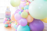 Clusters of pastel balloons in pink, purple, yellow, green, and blue brighten the room. In the background is a large cake topped with Anagram/MD’s 28" Holographic SuperShape Pearlized Birthday Foil Balloon.
