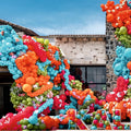 Des grappes de ballons en latex Kalisan de 45 cm, de couleur bleu aqua standard (25 par sachet), décorent un bâtiment en pierre ; de grandes lettres multicolores formées par des ballons épellent « KAI » à l'entrée, sous un ciel bleu.