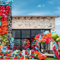 Un bâtiment en pierre est décoré de ballons en latex Kalisan de 12,7 cm (5 pouces) de couleur vert matcha standard, formant des arches et des grappes festives. De grandes baies vitrées reflètent le ciel et la verdure, mettant en valeur cette célébration animée en plein air.