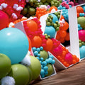 De grandes lettres décoratives blanches sont entourées et garnies de ballons festifs en latex Kalisan de 30 cm, de couleur orange vif standard, apportant une touche de couleur ludique à la décoration posée sur le parquet.