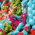 Une composition éclatante composée de grappes de ballons en latex de 30 cm aux couleurs bleu, orange, rose et vert matcha standard « Kalisan », rehaussée d'un grand papillon rouge en centre et de petites touches florales blanches disséminées parmi les ballons.
