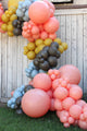 Una llamativa guirnalda de globos, elaborada con globos de látex de diseño «Poppy» de Tuftex de 17″ (50 unidades por bolsa) en colores rosa, amarillo, azul y marrón, está colocada frente a una pared rústica de madera blanca, con césped verde a sus pies.