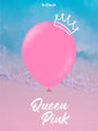 A large heart-shaped 12″ Kalisan latex balloon in Queen Pink, featuring a crown illustration, floats above a beach with blue waves. The words “Queen Pink” are written in white script at the bottom.