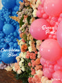 A vibrant balloon display features blue balloons on the left labeled “Caribbean Blue,” and pink balloons on the right labeled “12\” Kalisan Latex Heart Balloons Standard Queen Pink (25 Per Bag)“ from Kalisan, with pastel flowers arranged in the center. The floor is brown, and the Kalisan logo appears at the bottom right.