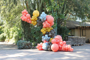 Una decoración festiva al aire libre en la que destacan globos de látex «Poppy Tuftex» de 24″ de la marca Tuftex, que forman un arreglo de diseño con globos de colores rosa, amarillo, azul y marrón junto a exuberantes plantas verdes, árboles y una estructura rústica de madera.